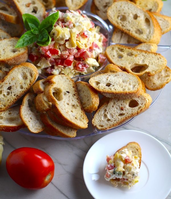 Bruschetta Topping surrounded by crostini on a platter with one crostini topped on a plate. It combines fresh tomatoes, artichoke hearts, garlic, basil, olive oil, and GOAT CHEESE!  Top on toasty garlic crostini and you get fresh bright flavors with both crunch and creamy in every bite.