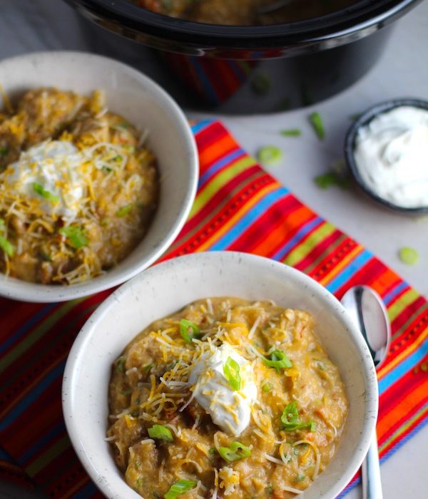 2 Bowls of Chipotle Chicken Enchilada Stew with sour cream and scallion slices as garnish and slow cooker pot in background. It's one of my Prepped Freezer Meal recipes and it's creamy, cozy, hearty, cheesy, and slightly spicy. All ingredients freeze raw, thaw, then cook in the slow cooker or in the oven. It's a stew because it's thick and hearty with bites of chicken, carrots, onion, chipotle peppers, cumin, garlic and tortilla chips that melt down to thicken this delicious stew.