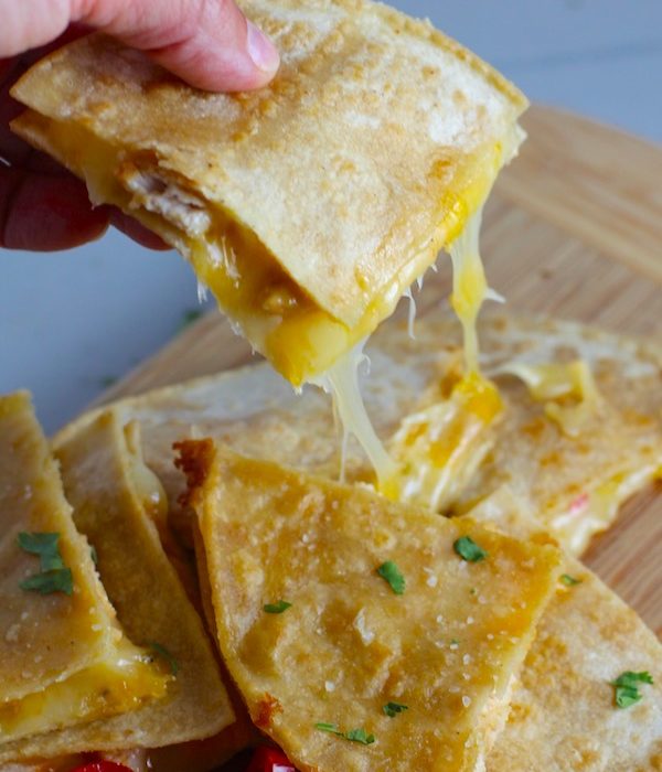 Hand holding Chicken Corn Tortilla Quesadilla triangle with melty cheese pulling. They have crispy edges, a soft gooey center, and are mouthwatering flavor. They have hearty shredded chicken flavored with warm and smoky Mexican spices. Then shredded Pepper Jack cheese and Cheddar is layered to get melty and oozy and delicious. It's an irresistible new easy dinner or appetizer idea.