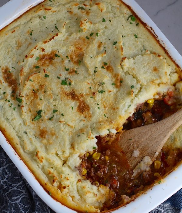 Spatula in Cottage Pie in casserole dish. This recipe has Spanish Chorizo and ground beef cooked in a rich and savory gravy with veggies and herbs.  Creamy mashed potatoes sit on top with manchego, parmesan, and garlic. #dinnerideas #cottagepie #shepherdspie