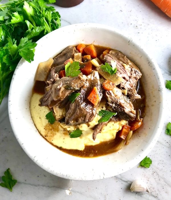 Cooked short ribs on top of creamy polenta in a bowl with sauce, carrots, and onions for flanken short ribs recipe slow cooker. Bowl sitting on counter.