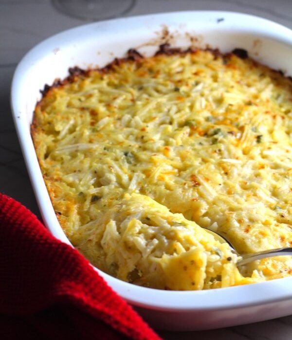 Spoon scooping Cheesy Hash Brown Casserole in casserole dish on counter with red towel. This recipe does not disappoint! It's warm, creamy, and full of flavor! And it's the perfect side dish for your holiday dinner.