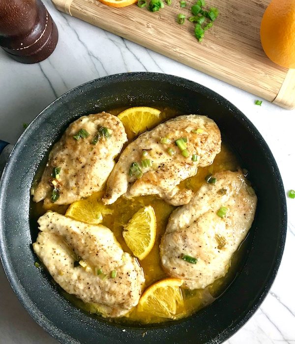 Orange Butter Sauce Chicken with orange slices and parsley garnish on top in skillet on counter with wood cutting board in background with sliced oranges and parsley.