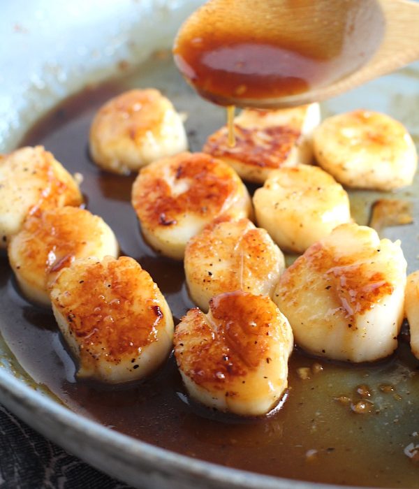 Wood spoon drizzling glaze on Glazed Pan Seared Scallops with Garlic and Honey in a pan with spatula.