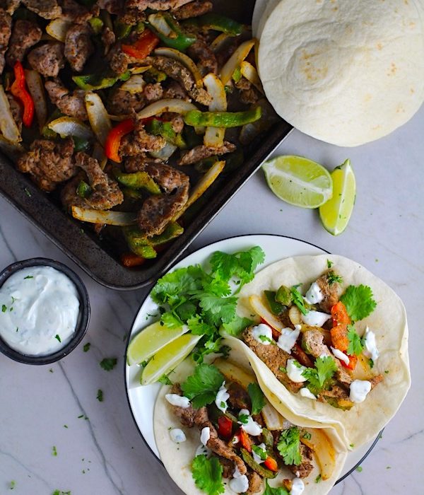 Sheet Pan Pork Fajitas Recipe with red pepper, green pepper, and onion, in flour tortillas on a plate. Topped with Cilantro Lime Crema, fresh cilantro leaves, and lime wedges. Sheet pan with pork, peppers, and onions in background along with crema and tortillas