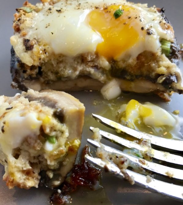 Portabella Mushroom Egg Nests on plate with bite cut out and runny yoke and fork on plate. Portabella mushrooms are filled with garlic, scallion, breadcrumb & Manchego cheese stuffing!  Then an egg is baked nestled in the center of all of this goodness.