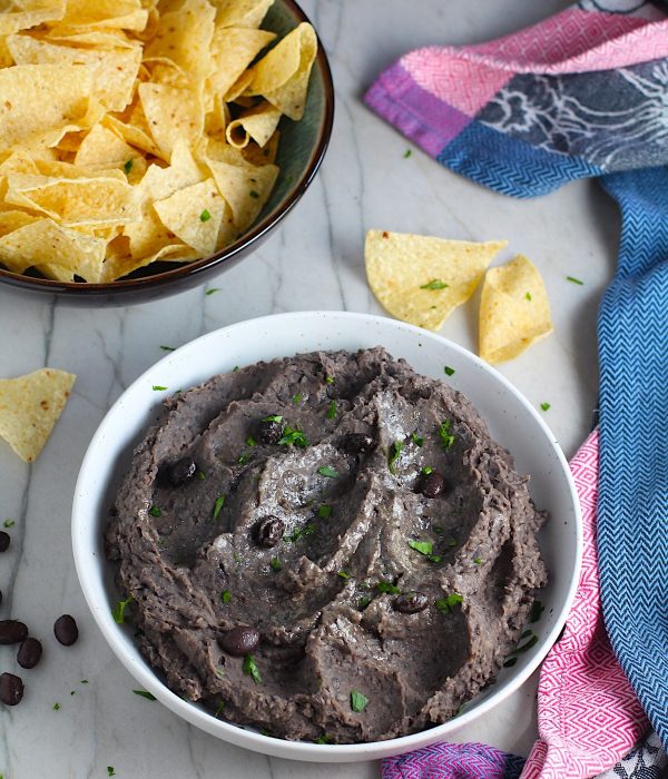 Creamy, rich, smokey, and delicious refried black beans recipe in a bowl with a few black beans and chopped cilantro sprinkled on top. On the counter are tortilla chips in a bowl.