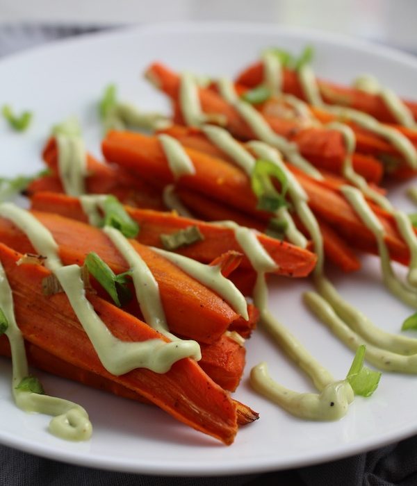 Close up of roasted carrots on plate with avocado crema drizzled on top with scallion slices. Roasted Carrots with smoky cumin and scallions, then topped with Avocado Crema are such a delicious and quick side dish for dinner.  Roasting brings out the natural sugar in the carrots, so they get a sweet caramelization with the salty and smoky flavors. The Avocado crema gives creamy and silky balance. 