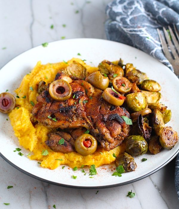 Sheet Pan Spanish Chicken thighs on a plate with Butternut Squash Mash, Olives, and Brussels Sprouts! It's warm, hearty, and smoky.