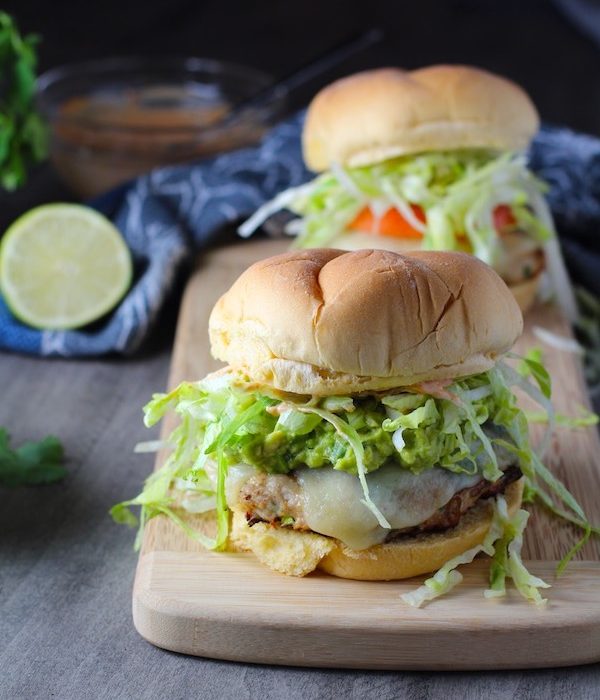 Two Taco Chicken Burgers on wood platter with smokey taco seasonings, cilantro, and scallions. Then on top are melty cheese, guacamole, lettuce, tomato, and chipotle mayonnaise!