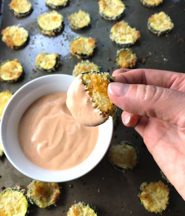 Breaded Zucchini Chips on pan with Sriracha dipping sauce in a bowl on pan. Hand dipping one zucchini chip into sauce.