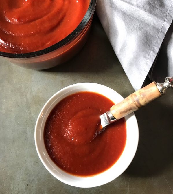 Best Sugar Free Ketchup in a white serving bowl with spreading knife and large bowl of ketchup in background.